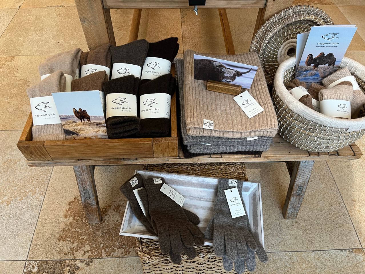 Ein Holzregal zeigt verschiedene Socken und Handschuhe. Die Socken sind in verschiedenen Farben und Größen und ordentlich in Körben angeordnet. In der Nähe sind einige gefaltete Decken. Ein Holzschild mit einem Bild eines Stiers ist auf einer Decke platziert.