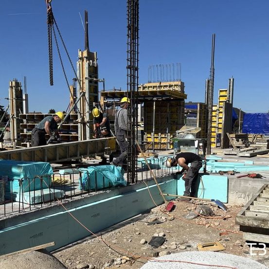 Bauarbeiter arbeiten auf einer Baustelle. Sie tragen Schutzhelme und arbeiten am Fundament. Eine Maschine hebt schwere Materialien. Hinter ihnen befinden sich weitere Gebäude und Strukturen.