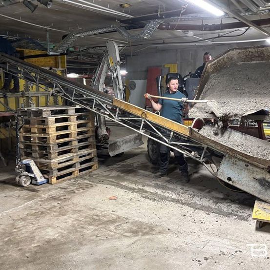 Ein Arbeiter in einer Fabrik benutzt eine Schaufel, um Material von einem Förderband in einen LKW zu schieben. Ein Stapel Holzpaletten und ein Palettenhubwagen sind in der Nähe.