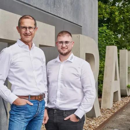 Zwei Männer in weißen Hemden und Brillen stehen vor einem großen 'BRA'-Schild mit Bäumen im Hintergrund.