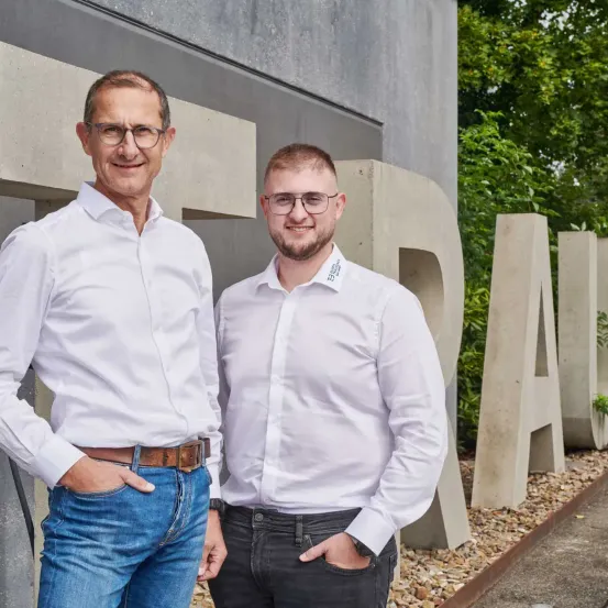 Zwei Männer in weißen Hemden und Brillen stehen vor einem großen 'BRA'-Schild mit Bäumen im Hintergrund.