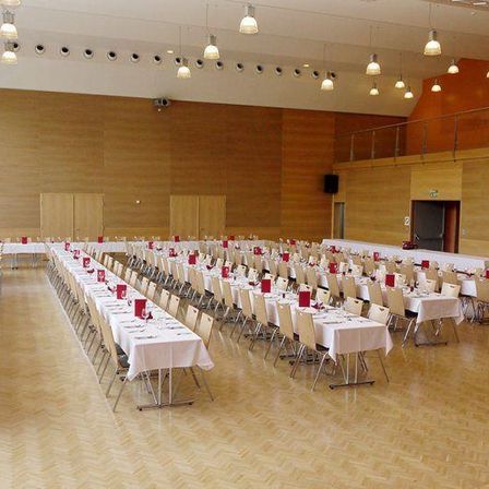 Ein Bankettsaal ist mit Tischen, Stühlen und weißen Tischdecken eingerichtet. Der Raum hat Holzwände, eine Galerie und einen Lautsprecher.