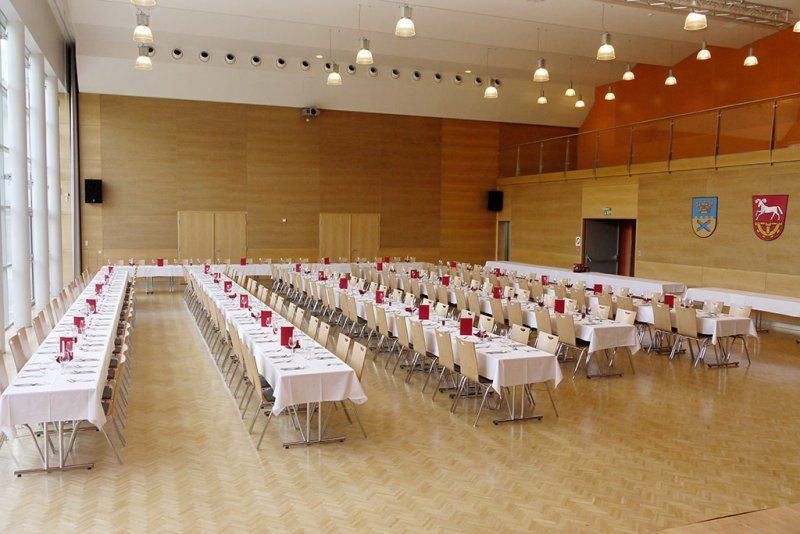 Ein Bankettsaal ist mit Tischen, Stühlen und weißen Tischdecken eingerichtet. Der Raum hat Holzwände, eine Galerie und einen Lautsprecher.