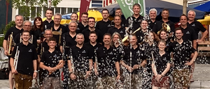 Eine Gruppe von Musikern feiert vor einem Gebäude, hält Musikinstrumente in den Händen, mit Konfetti und einer grünen Fahne im Hintergrund.