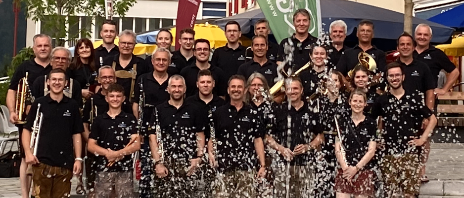 Eine Gruppe von Musikern feiert vor einem Gebäude, hält Musikinstrumente in den Händen, mit Konfetti und einer grünen Fahne im Hintergrund.