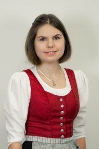 Eine Frau mit kurzen Haaren lächelt für ein Foto. Sie trägt eine rote Weste mit weißen Knöpfen und ein weißes Oberteil. Sie hat eine Halskette mit einem Anhänger.
