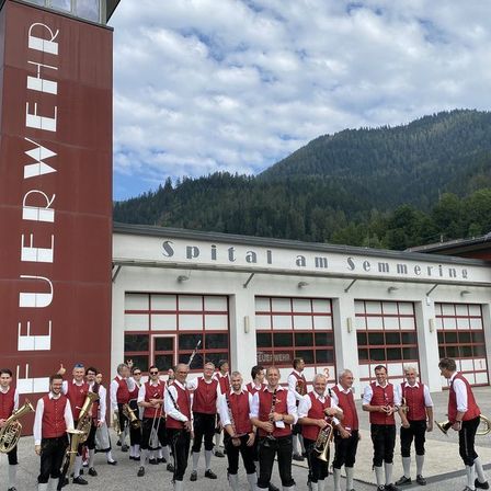 Eine Gruppe von Musikern in traditioneller Kleidung steht vor dem Fluerwehr-Gebäude mit Bergen im Hintergrund.