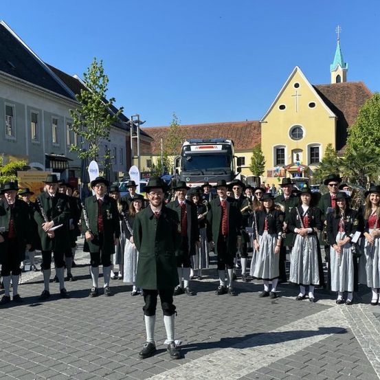 Eine Gruppe von Musikern in traditioneller Kleidung steht in einer Reihe, mit einem LKW und einer Kirche im Hintergrund.