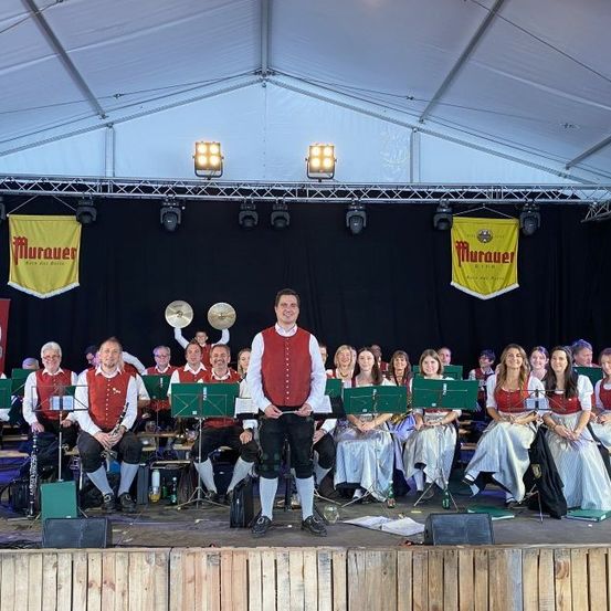 Ein Blasorchester auf der Bühne mit Musikinstrumenten, Fahnen und Scheinwerfern. Die Mitglieder tragen traditionelle Kleidung, mit einer Mischung aus sitzenden und stehenden Positionen.