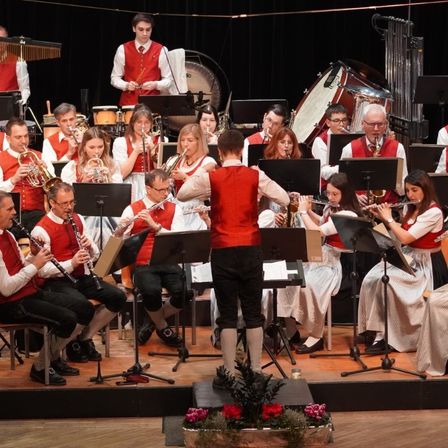 Ein großes Orchester tritt auf der Bühne auf und besteht aus Musikern, die verschiedene Instrumente spielen. Einige sitzen auf Stühlen, andere stehen. Der Dirigent steht vorne und leitet die Aufführung.