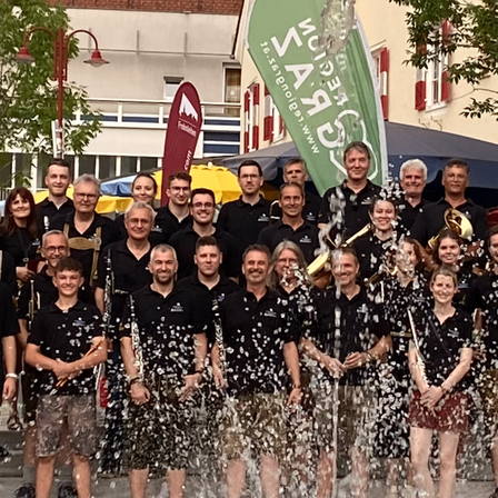 Eine Gruppe von Menschen in Schwarz feiert vor einem grünen Banner mit Wasserspritzern. Im Hintergrund befinden sich Gebäude, Bäume und ein rotes Banner.
