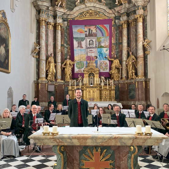 Ein Chor in formeller Kleidung tritt in einer Kirche auf, mit Musikern, die verschiedene Instrumente spielen. Ein Mann steht vor einem Podium, und dahinter ist ein Banner angebracht.