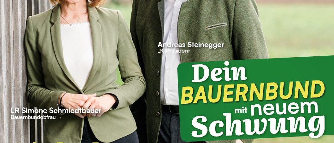 Ein Paar in formeller Kleidung steht lächelnd für ein Foto. Die Frau trägt einen grünen Blazer und eine Halskette, und der Mann trägt eine Brille und einen grünen Anzug. Sie stehen neben einem großen grünen Banner mit Text auf Deutsch und Englisch. Das Banner lautet 'Einladung zum Landesbauerntag'.