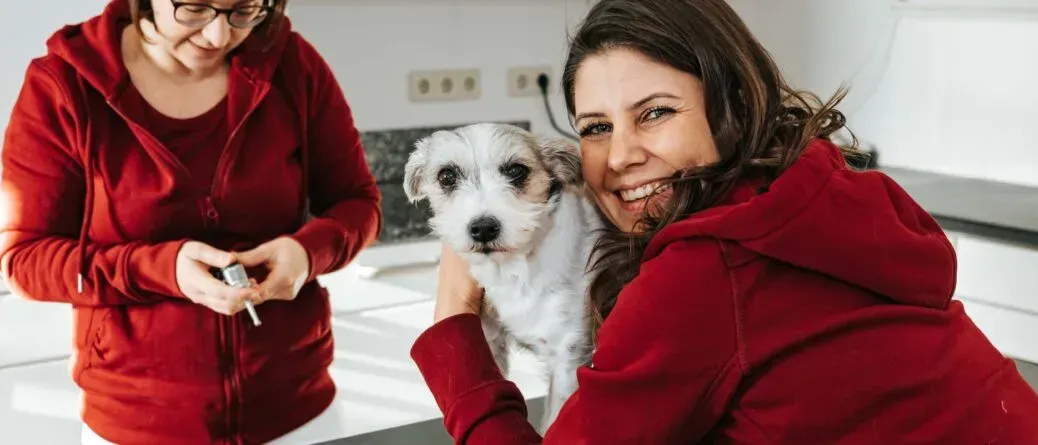 Zwei Frauen in roten Jacken sind in einer Küche, eine hält einen Hund und die andere einen Stift.