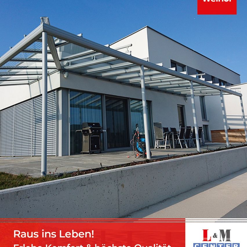 Ein Bild eines modernen Hauses mit einem Terrassenbereich, der eine Glasüberdachung, einen Grill und einen Tisch mit Stühlen aufweist. Es gibt einen Wasserzeichen, der 'Raum ins Leben!' in rotem Schriftzug zeigt.
