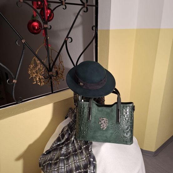 Eine grüne Handtasche mit einem Löwenemblem steht auf einer weißen Oberfläche, begleitet von einem grünen Hut. Dahinter hält eine dekorative Metallstruktur rote Ornamente.
