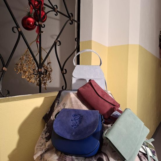 Mehrere Handtaschen in verschiedenen Farben sind auf einem Regal ausgestellt, mit einer gelben und weißen Wand im Hintergrund. Ein schwarzer Metallrahmen hält rote Weihnachtskugeln.