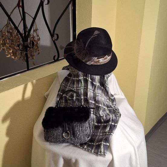 Ein schwarzer Hut mit einer Feder und einem Schal liegt auf einem Tisch mit einer Tasche und einem weißen Tuch. Ein Metallgitter befindet sich hinter dem Tisch.