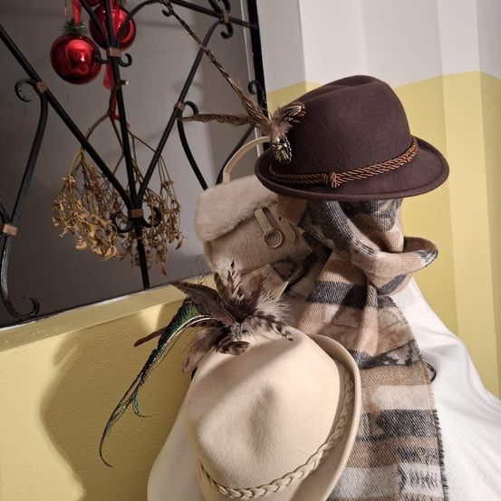 Ein Raum mit gelber Wand zeigt zwei Hüte und einen Schal. Die Hüte haben Federn und einer hat eine dekorative Brosche. Der Schal ist gestreift und hängt neben den Hüten.