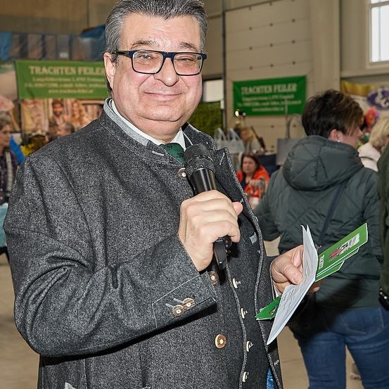 Ein Mann mit Brille und einem grauen Mantel hält ein Mikrofon und spricht bei einer Veranstaltung. Er hat ein grünes Büchlein in der Hand, und hinter ihm steht eine weitere Person. Im Hintergrund zeigt ein Banner die Worte 'Trachten Feier'.