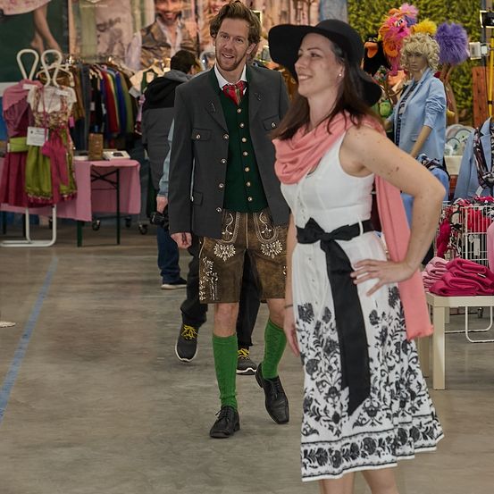 Ein Mann in traditioneller bayerischer Kleidung und eine Frau in einem weißen Kleid mit einem rosa Schal stehen in einem Bekleidungsgeschäft.