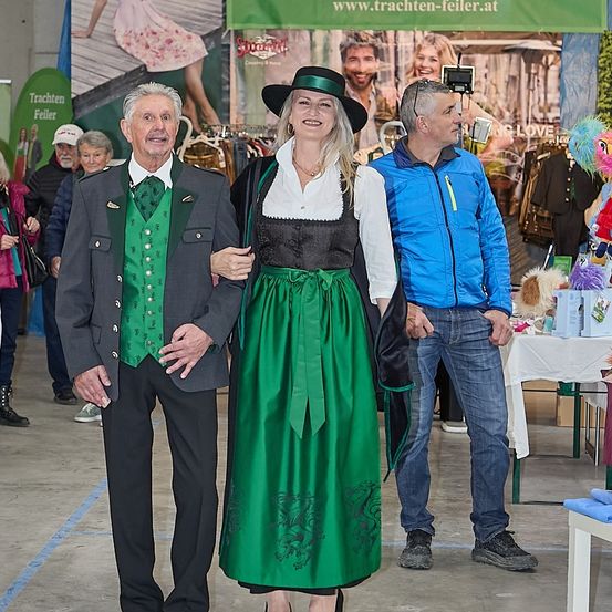 Ein älterer Mann und eine Frau in traditioneller bayerischer Tracht stehen neben einem Mann in Freizeitkleidung. Sie befinden sich in einer Halle mit einem Hintergrund eines Geschäfts.