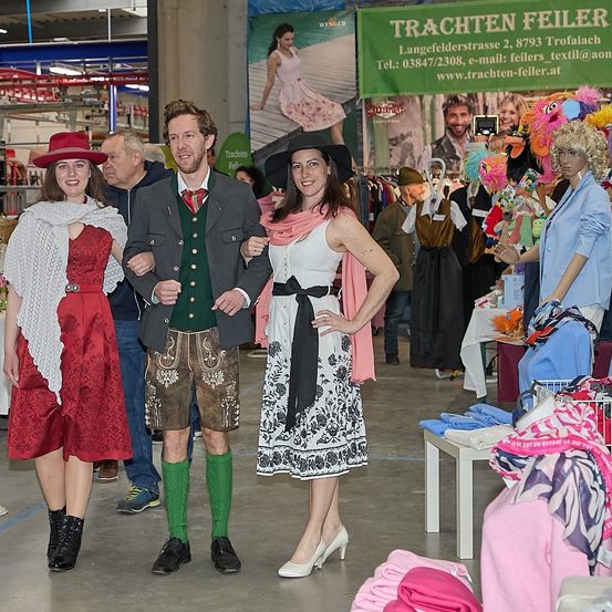 Drei Menschen in traditioneller Kleidung stehen in einem Bekleidungsgeschäft und lächeln. Ein Mann in traditioneller Kleidung steht zwischen zwei Frauen. Ein Banner mit einer Frau und dem Text Trachten Feier ist im Hintergrund.