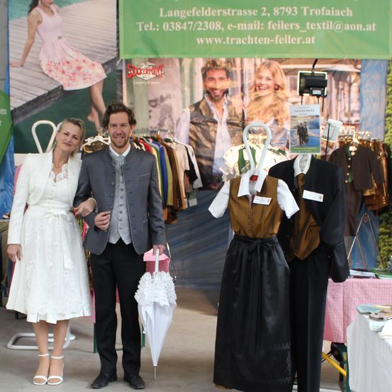 Ein Paar steht neben traditioneller Kleidung, die ausgestellt ist, mit einem Hintergrund von einem lächelnden Mann und einer Frau. Der Mann hält einen weißen Schirm.
