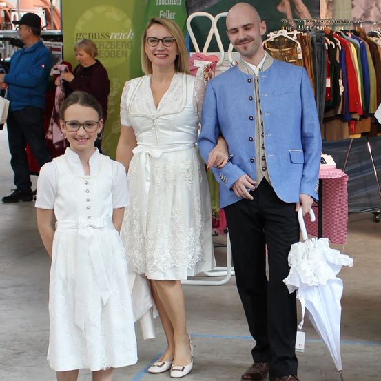 Eine Frau und ein Mann in traditioneller bayerischer Kleidung stehen mit einem Mädchen in einem weißen Kleid. Sie lächeln und stehen vor einem Tisch mit Kleidung.