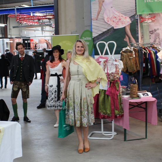 Eine Frau steht in einem Einkaufszentrum mit zwei anderen Frauen, eine trägt einen Hut. Ein Tisch mit Kleidung und Taschen steht hinter ihnen.