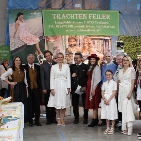 Gruppenfoto bei einer Veranstaltung mit einem grünen Banner mit den Details von Trachten Feierer. Mehrere Erwachsene und Kinder in traditioneller Kleidung posieren vor einer Kulisse mit einer Frau und einem Mann.
