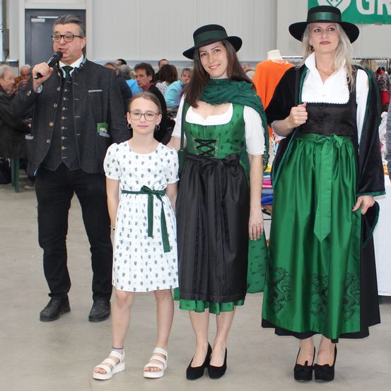 Ein Mann mit Mikrofon steht mit drei Frauen in traditioneller deutscher Kleidung, vermutlich bei einer kulturellen Veranstaltung.