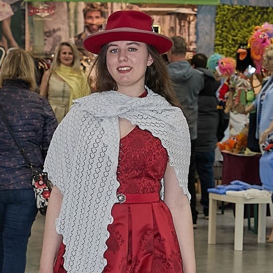 Eine Frau in einem roten Kleid und einem weißen Spitzenschal geht durch einen Flur, mit anderen Menschen und Gegenständen im Hintergrund.
