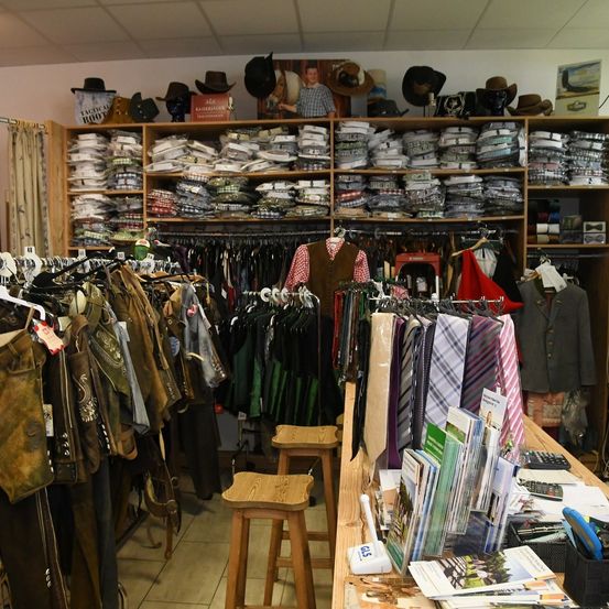 Ein Bekleidungsgeschäft mit verschiedenen Artikeln wie Krawatten, Hüten und Westen. Regale mit ordentlich gefalteten Hemden und anderen Kleidungsstücken. Holzstühle und Tische mit Broschüren und einem Taschenrechner.