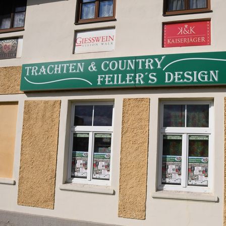 Die Schaufensterfront von Trachten & Country Feier's Design, einem Geschäft mit Fenstern, die Werbematerialien präsentieren. Über dem Ladenschild befinden sich Schilder für GIESSWEIN Vision Walk und K&K Kaiserjäger.