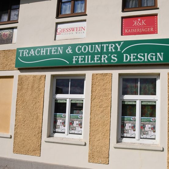 Die Schaufensterfront von Trachten & Country Feier's Design, einem Geschäft mit Fenstern, die Werbematerialien präsentieren. Über dem Ladenschild befinden sich Schilder für GIESSWEIN Vision Walk und K&K Kaiserjäger.