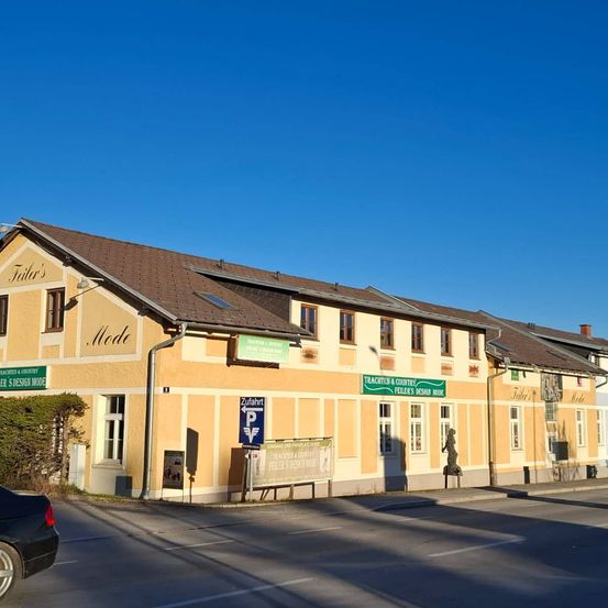 Ein Gebäude mit einem braunen Dach, das mehrere Schilder aufweist, darunter eines für Trachettes & Country und ein anderes für Schneider's Design Mode. Ein schwarzes Auto ist am Straßenrand geparkt.