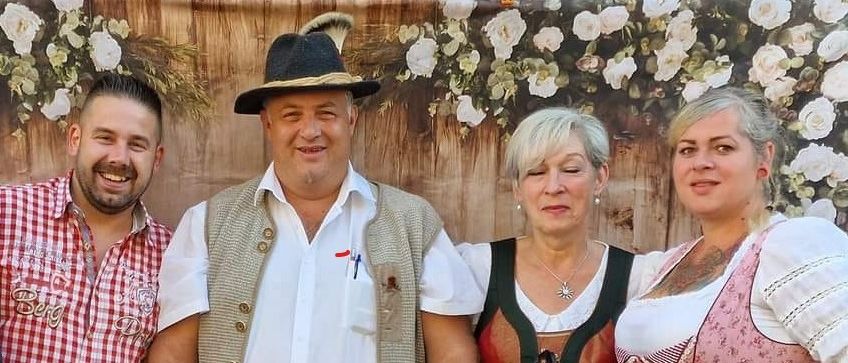 Vier Personen in traditioneller bayerischer Kleidung stehen vor einem rustikalen Holzhintergrund. Zwei Männer und zwei Frauen, lächelnd, tragen unterschiedliche traditionelle Kostüme und Accessoires.