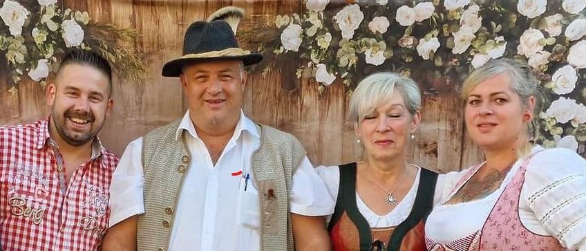 Vier Personen posieren für ein Foto vor einem Hintergrund aus Blumen und Holz. Die beiden Männer tragen traditionelle bayerische Kleidung, und die beiden Frauen tragen traditionelle deutsche Kleidung.