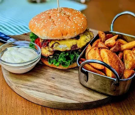 Ein Holzschneidebrett mit einem Hamburger, einer Beilage Pommes und einer Schüssel Mayonnaise. Der Burger hat ein Sesambrötchen, Salat, Tomate und eine Scheibe mit Käse. Die Pommes sind goldbraun.