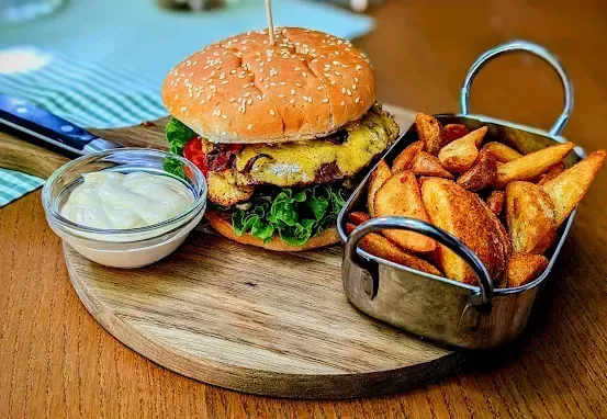 Ein Holzschneidebrett mit einem Hamburger, einer Beilage Pommes und einer Schüssel Mayonnaise. Der Burger hat ein Sesambrötchen, Salat, Tomate und eine Scheibe mit Käse. Die Pommes sind goldbraun.