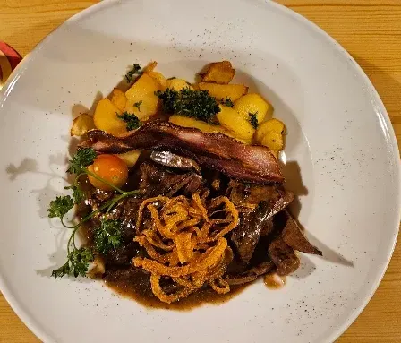 Ein weißer Teller mit einem herzhaften Gericht aus Fleisch, Kartoffeln und Zwiebelringen, begleitet von einem Glas Bier auf einem Holztisch.