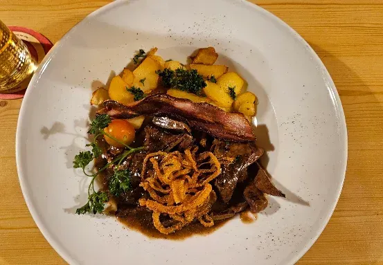 Ein weißer Teller mit einem herzhaften Gericht aus Fleisch, Kartoffeln und Zwiebelringen, begleitet von einem Glas Bier auf einem Holztisch.