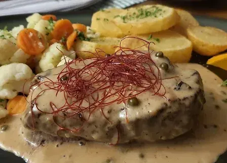 Ein Teller mit einem in Sauce getauchten Steak, garniert mit roten Chilifäden, begleitet von Kartoffelpüree, Blumenkohl, Karotten und Brokkoli, mit einer Zitronenscheibe auf der Seite.