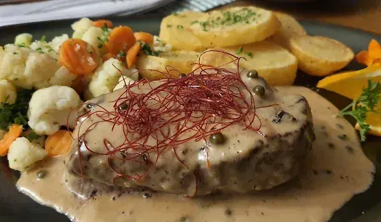 Ein Teller mit einem in Sauce getauchten Steak, garniert mit roten Chilifäden, begleitet von Kartoffelpüree, Blumenkohl, Karotten und Brokkoli, mit einer Zitronenscheibe auf der Seite.