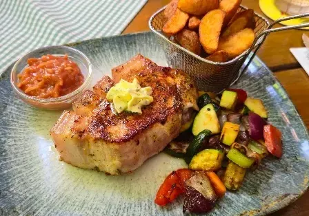 Ein Teller mit gegrilltem Schweinekotelett mit Gemüse und einer Beilage Pommes, begleitet von einer kleinen Schüssel Sauce und einem Korb Pommes.