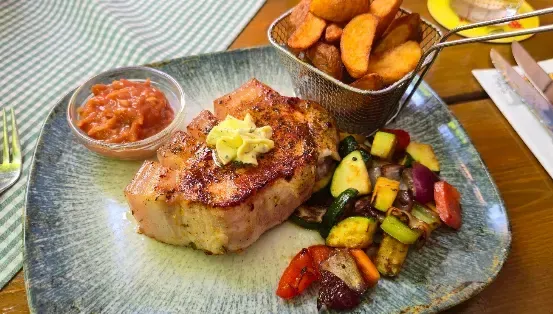 Ein Teller mit gegrilltem Schweinekotelett mit Gemüse und einer Beilage Pommes, begleitet von einer kleinen Schüssel Sauce und einem Korb Pommes.