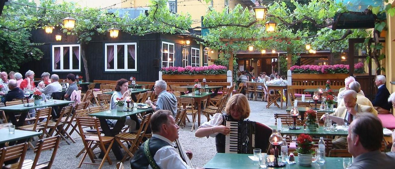 Ein Essbereich im Freien mit Menschen an Tischen. Der Ort ist mit Blumen geschmückt und hat ein hölzernes Gazebo. Eine Frau spielt Akkordeon, während der Mann Gitarre spielt.