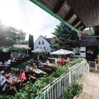 Ein Außenbereich mit Menschen, die um Tische in einem Garten sitzen, mit Sonnenschirmen und einer weißen Umzäunung. Ein weißes Haus mit Schornstein ist im Hintergrund.