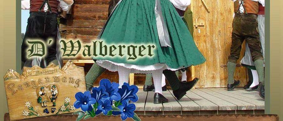 Eine Gruppe von Menschen in traditioneller bayerischer Kleidung tanzt auf einer Holzbühne. Im Vordergrund sind blaue Blumen zu sehen. Das Schild trägt den Namen 'D' Wallberger'.